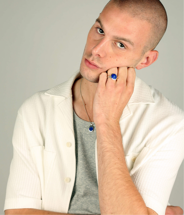 Man wearing a white shirt over a gray shirt with a blue enamelled skull ring on his finger and a similar necklace, against a plain background